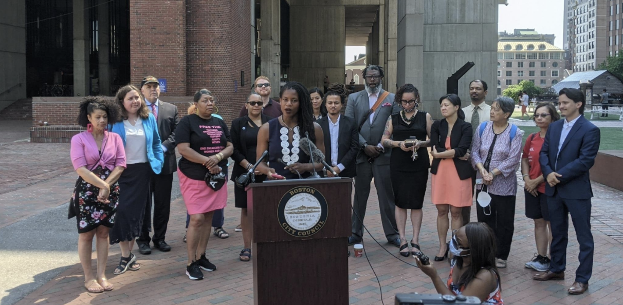 image of Councilor Lydia Edwards giving a press conference in front of Boston's City Hall, with a group of sixteen people standing in a semi-circle a few paces behind her; there is a person holding a recorder, crouched in the bottom, right of the photo, wearing a mask, and the dark wood podium Councilor Edwards is standing at has the official Boston City Council seal on the top portion