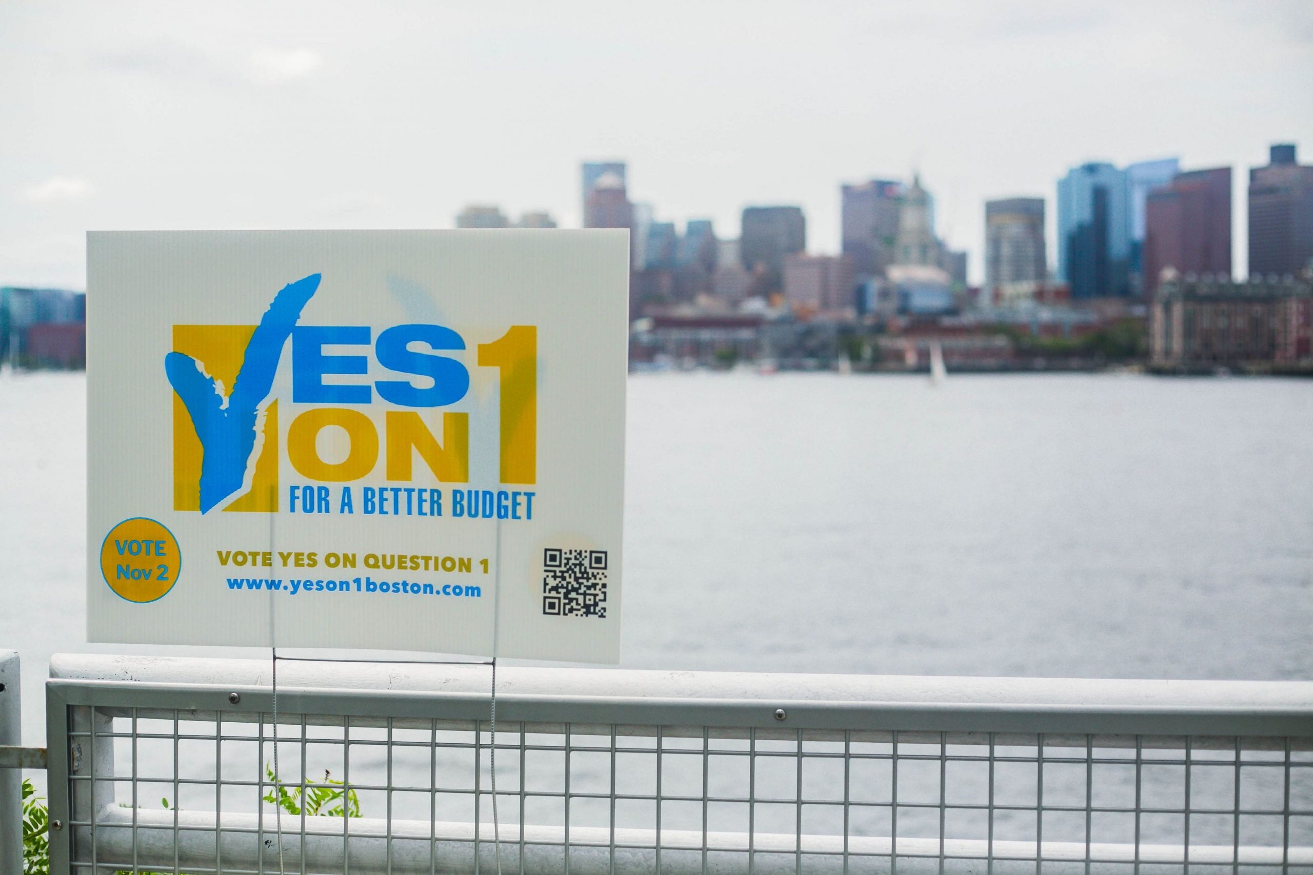a Yes on 1 sign in focus in front of a fence, in the background there is water and the Boston skyline