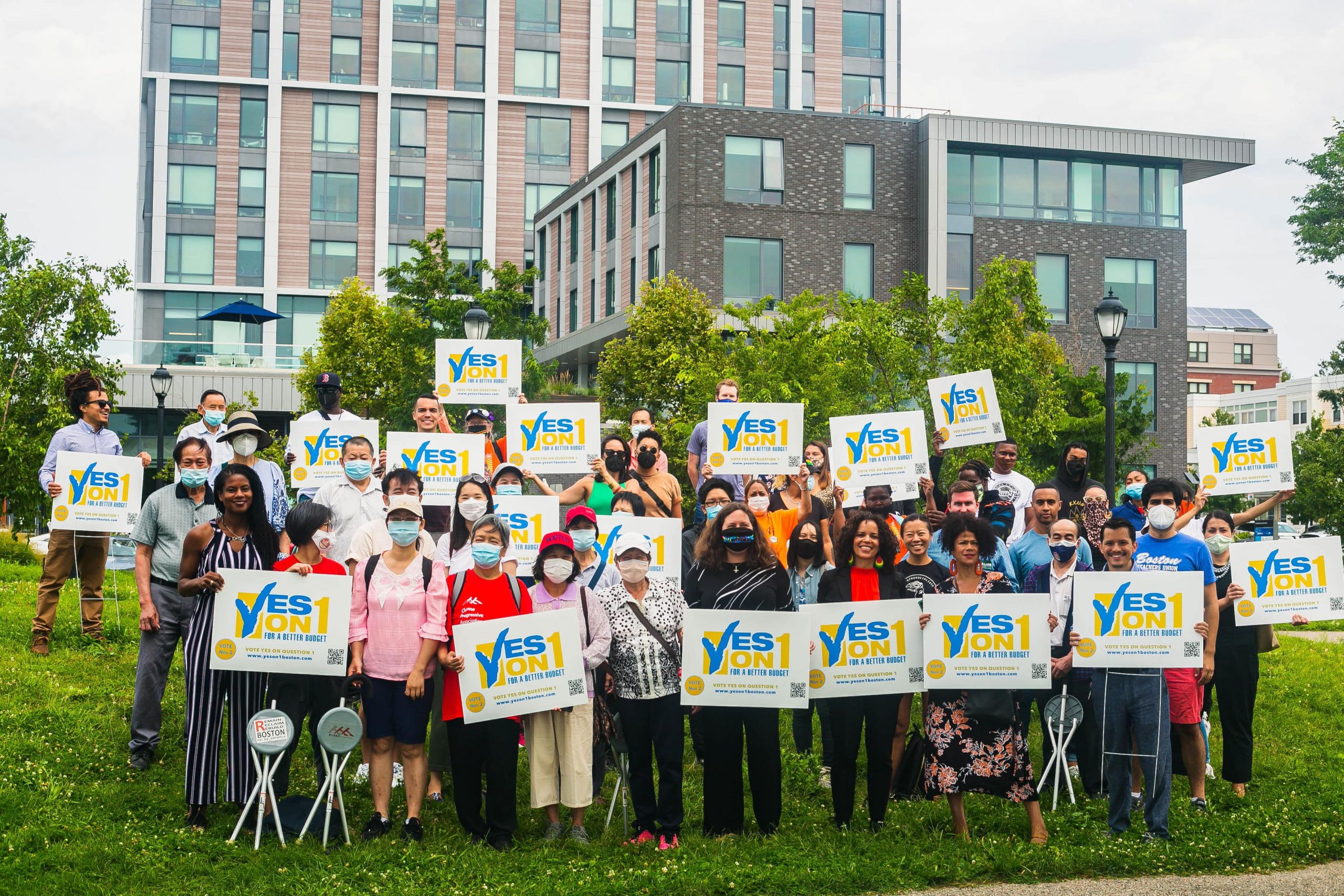 photo of volunteers on a green lawn holding yes on 1 signs, with building in the background